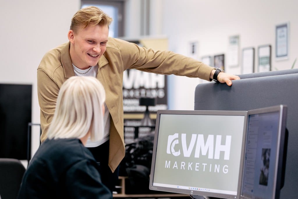 Hymyilevä mies seisoo VMH Marketingin toimistossa ja nojaa toimistotuolin selkänojaan samalla, kun hän keskustelee iloisen naisen kanssa, joka työskentelee tietokoneella. Tietokoneen näytöllä näkyy VMH Marketingin logo.
